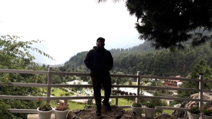 Tejas standing at a railing above a green valley ringed by pine forest and hills