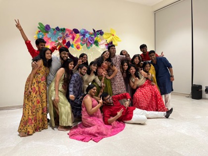 Around eighteen friends in festive Indian dress crowded together in front of a handmade paper flower wall