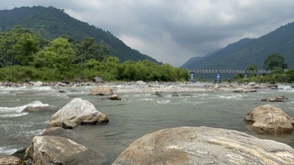 A wide river running over boulders between green hills, a bridge in the distance